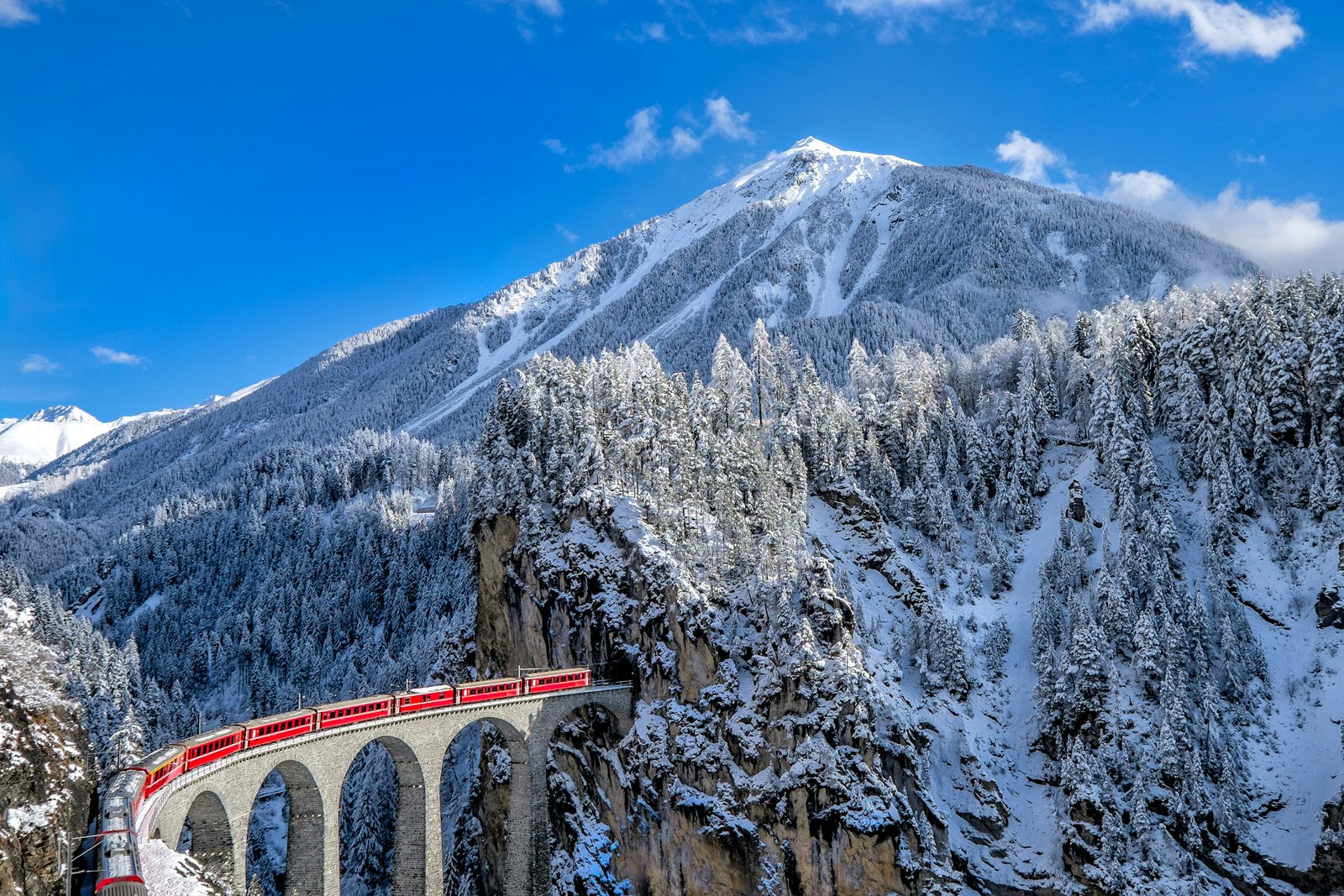 The bright red Glacier Express train cuts through rugged mountains covered in snow and trees under a clear blue sky